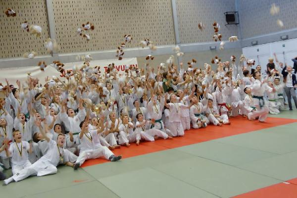 Groepsfoto van veruit alle judoka's die hun ontvangen knuffel de lucht ingooien.